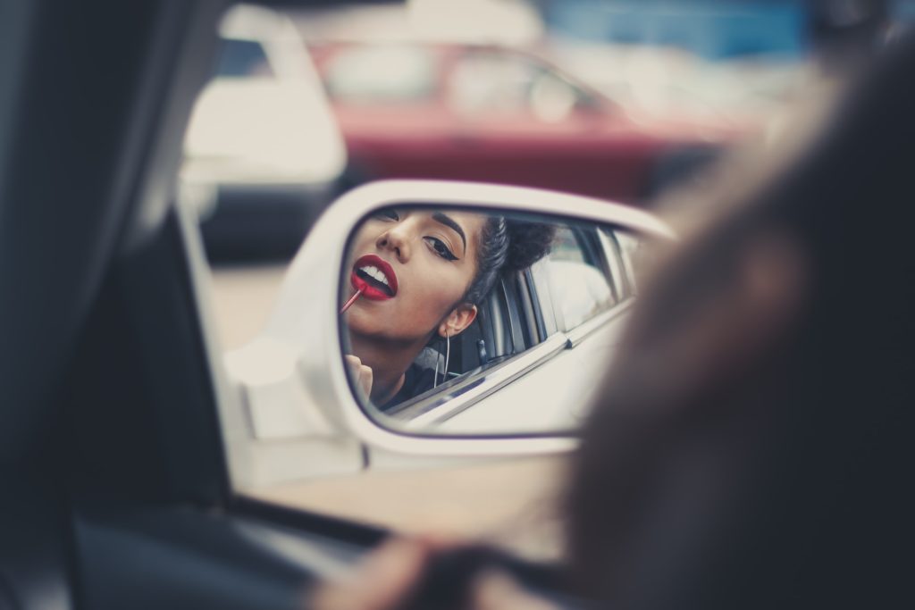 Woman putting on lipstick in the side mirror of her car.